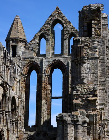 This architectural photograph captures the ruins of Whitby Abbey, a historic church located in Whitby, Yorkshire. Taken in the early afternoon during the spring season, the image shows the impressive stone structure of the abbey silhouetted against a bright blue sky. Prominently visible through the pointed arches and gothic windows is the moon, adding a distinctive element to the scene. Whitby Abbey is a well-known landmark in Yorkshire, and its ancient, weathered walls and intricate details are clearly showcased in this architectural shot. The photograph highlights both the cultural heritage of Whitby and the iconic church ruins that overlook the North Sea, reflecting the enduring beauty and significance of Whitby Abbey in Yorkshire.